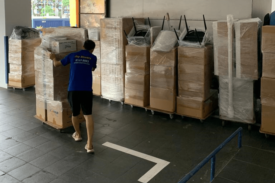 Person in a blue shirt pushes a covered pallet in a storage area. Multiple wrapped items, including chairs and boxes, are stacked against the wall.