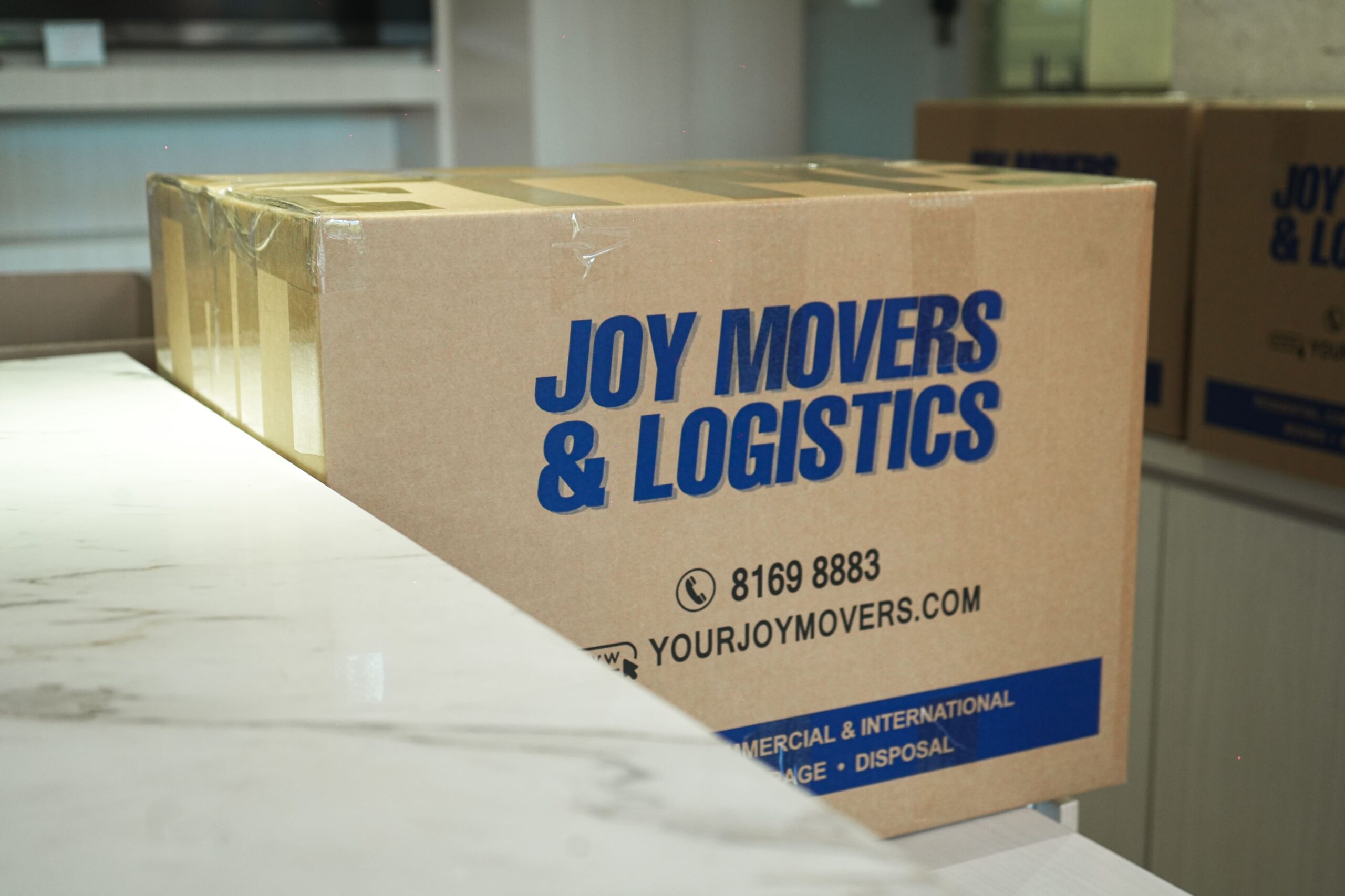 A cardboard box with the label "Joy Movers & Logistics" sits on a countertop. More similar boxes are in the background, highlighting their comprehensive house moving services.