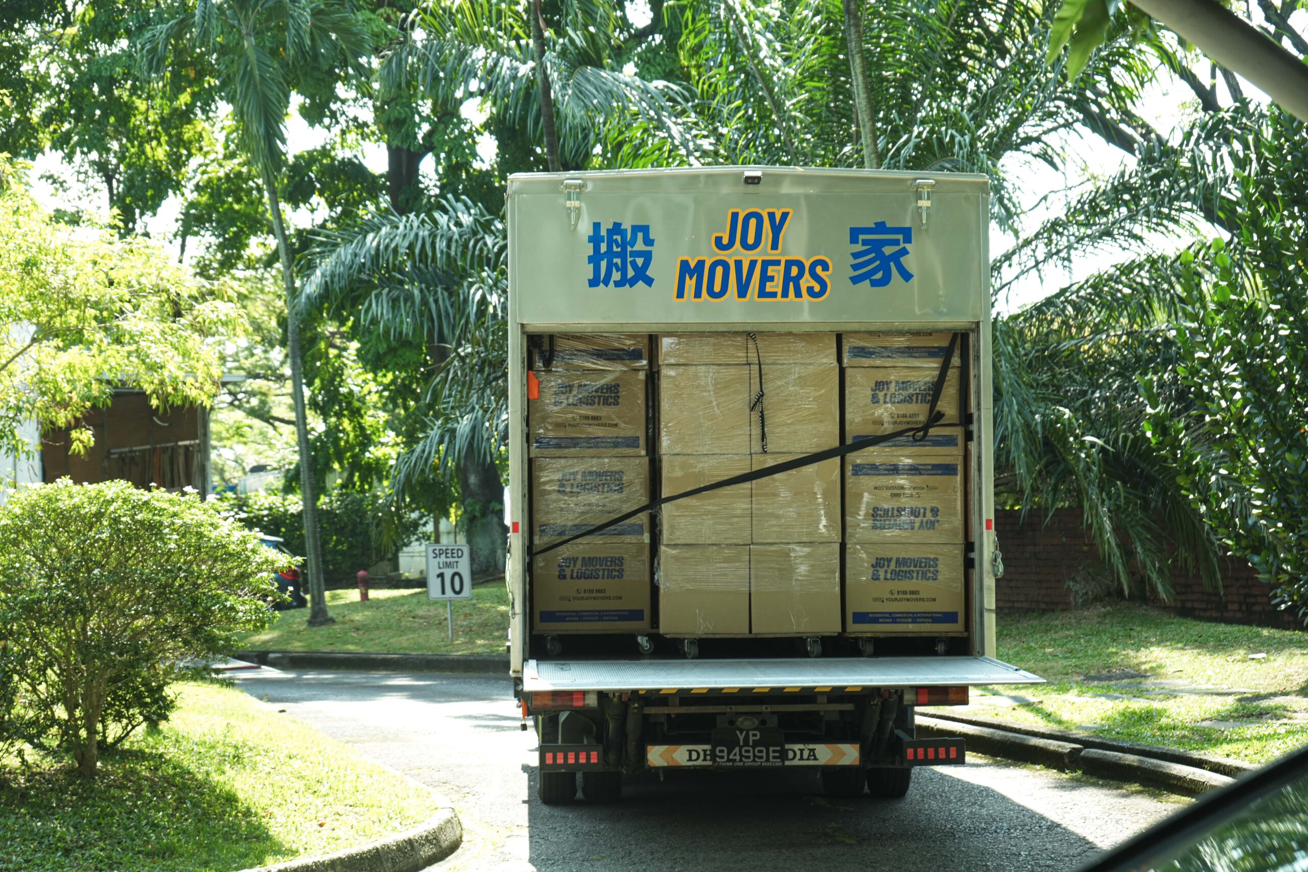 A moving truck with the words "JOY MOVERS" and Chinese characters printed on the back is loaded with stacked cardboard boxes.